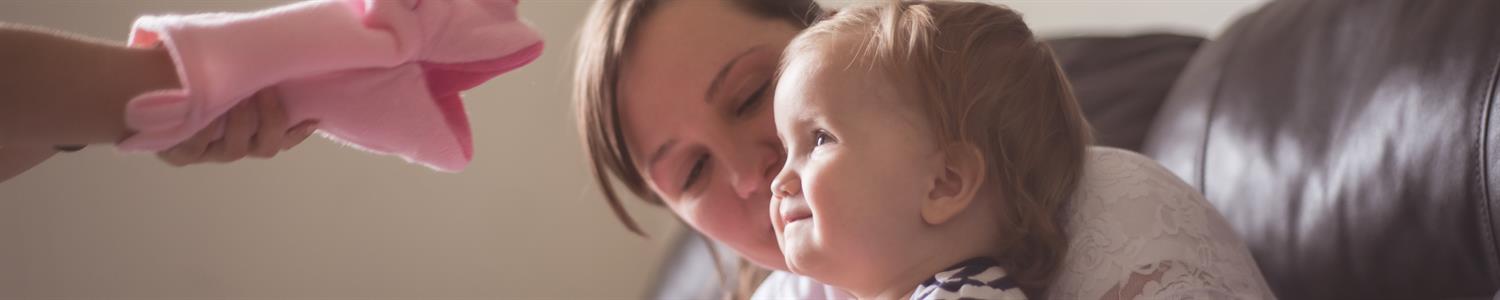 Image of a young mom holding a baby laughing at a puppet.