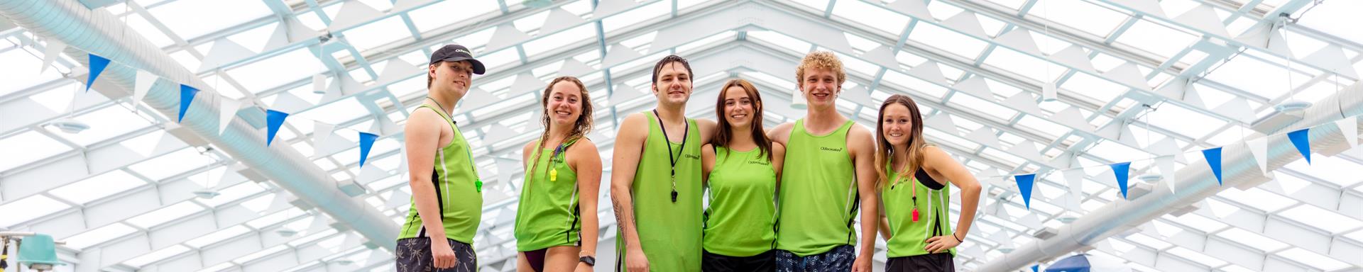 Four lifeguards in uniform sitting on the pool deck facing the water looking back at the camera.