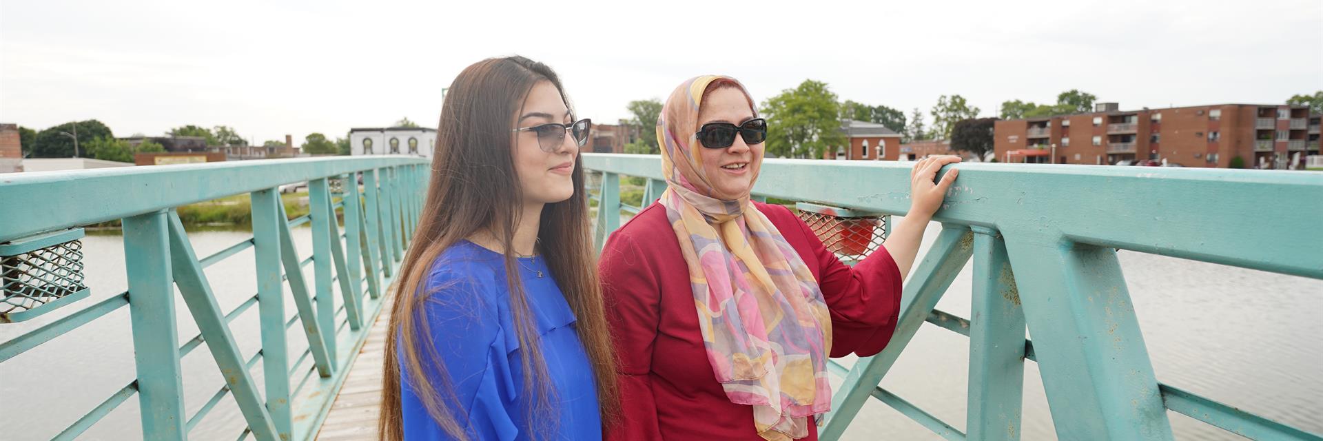 Two women on a teal walking bridge over a river.