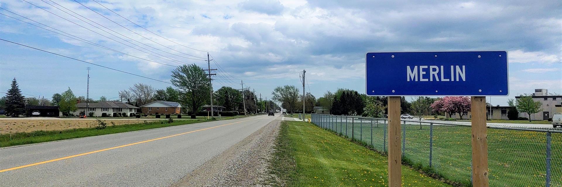 Merlin road sign with a public school in the background and houses on the opposite side of the road.