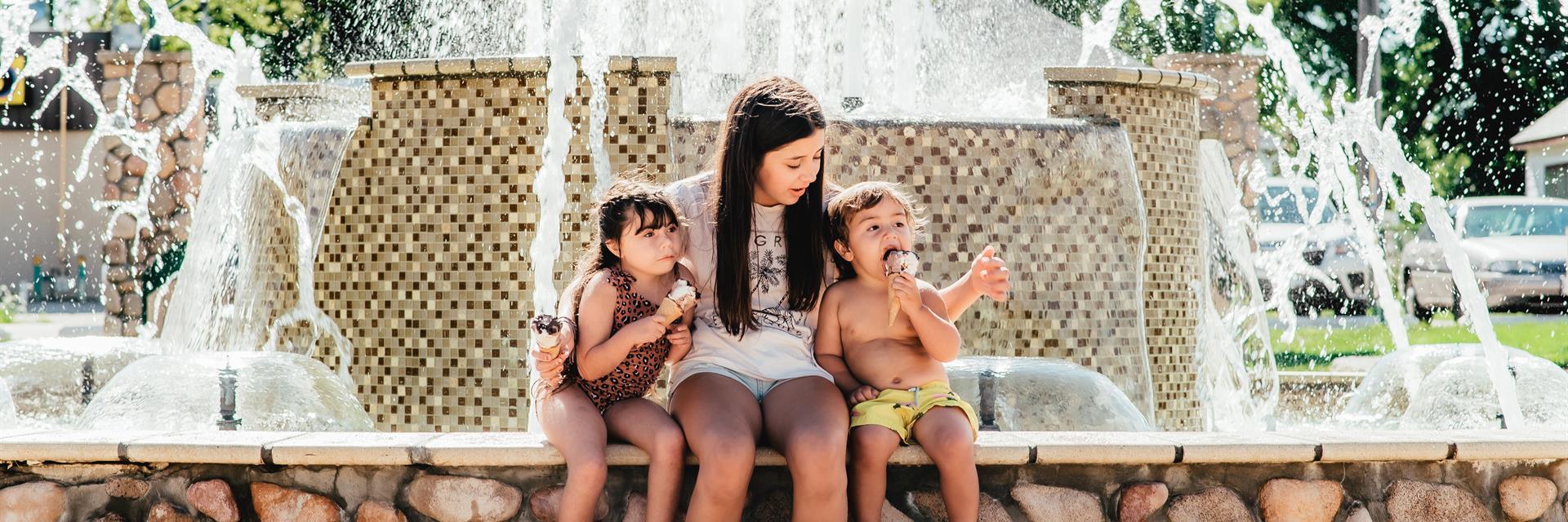 Mother and two children sitting on the side of a large fountain.